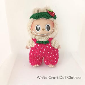 Op de afbeelding: Een pop gekleed in een aardbeienkostuum. De pop heeft een crèmekleurige harige capuchon met een groene hoed en een rode aardbei. Ze draagt een rode tuinbroek met witte stippen. De tekst "White Craft Doll Clothes" staat onderaan.
