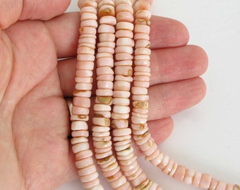 Op de afbeelding: Een streng roze en witte schelp kralen. De kralen zijn klein en rond en hebben een natuurlijke, organische uitstraling. De kralen zijn geregen op een doorzichtige draad.
