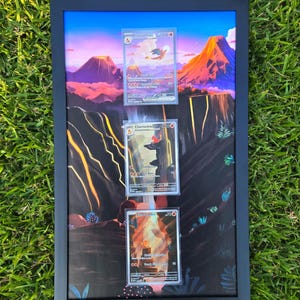 May include: A framed artwork featuring three Pokemon trading cards. The cards are displayed against a backdrop of a colorful landscape with mountains and a volcano. The frame is black, and the artwork is set on a green grassy surface.
