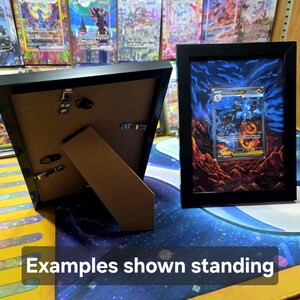 Puede incluir: Dos exhibidores de cartas coleccionables enmarcados en negro. Un marco est&aacute; abierto, mostrando la parte posterior con un soporte. El otro marco muestra una tarjeta colorida con un fondo azul y naranja. El texto "Examples shown standing" es visible.