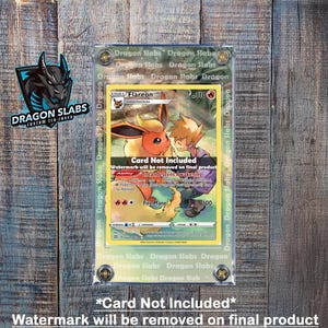 May include: A Pokemon trading card in a clear protective case. The card shows a Flareon illustration with a trainer. The case is labelled "Dragon Slabs" and has the text "Card Not Included". The background is a wooden surface.