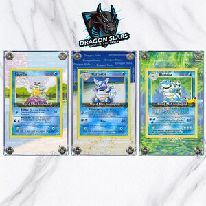 May include: Three Pokemon trading cards in protective cases. The cards feature Squirtle, Wartortle, and Blastoise, with blue and green backgrounds. The cases are clear with the "Dragon Slabs" logo visible.