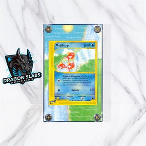 Peut inclure: Une carte Pokémon Magikarp classée dans un étui en acrylique transparent. La carte présente une bordure jaune et une illustration de Magikarp rouge. L'étui est étiqueté "Dragon Slabs Custom TCG Cases" avec un logo de dragon.