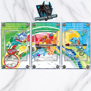 May include: Three clear display cases for trading cards, each featuring a different Pokémon character: Bulbasaur, Charmander, and Squirtle. The cases have a gold-colored border and the text "Dragon Slabs" is visible. The background is a marble pattern.