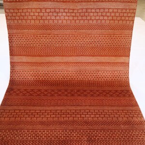 K&ouml;nnte beinhalten: Ein langer, schmaler Teppich mit einem sich wiederholenden geometrischen Muster in Orange- und Wei&szlig;t&ouml;nen. Der Teppich ist in der Mitte gefaltet und liegt auf einer wei&szlig;en Oberfl&auml;che.