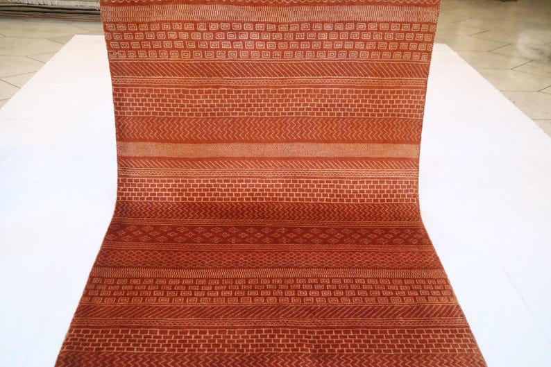 K&ouml;nnte beinhalten: Ein langer, schmaler Teppich mit einem sich wiederholenden geometrischen Muster in Orange- und Wei&szlig;t&ouml;nen. Der Teppich ist in der Mitte gefaltet und liegt auf einer wei&szlig;en Oberfl&auml;che.