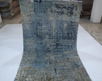 Puede incluir: Una alfombra rectangular con un diseño abstracto. La alfombra presenta una mezcla de tonos azules, grises y beige, creando una apariencia texturizada. El diseño tiene un aspecto desgastado y usado, con variaciones de color y patrón.