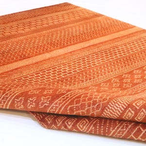 K&ouml;nnte beinhalten: Ein Nahaufnahme eines gefalteten orangefarbenen und wei&szlig;en Stoffes mit geometrischem Muster. Der Stoff ist weich und texturiert.