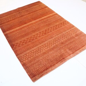 K&ouml;nnte beinhalten: Ein rostfarbener Teppich mit einem geometrischen Muster, das in den Stoff eingewebt ist. Der Teppich hat ein gestreiftes Design mit verschiedenen Mustern in jedem Streifen.