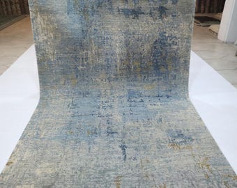Puede incluir: Una alfombra enrollada con un diseño abstracto y desgastado. La alfombra presenta una mezcla de tonos azules, grises y beige, creando una apariencia texturizada. La alfombra se exhibe sobre una superficie blanca.
