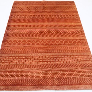 K&ouml;nnte beinhalten: Ein rostiger Teppich mit einem geometrischen Muster aus wei&szlig;en Linien und Formen. Der Teppich hat eine gewebte Textur und eine rechteckige Form.