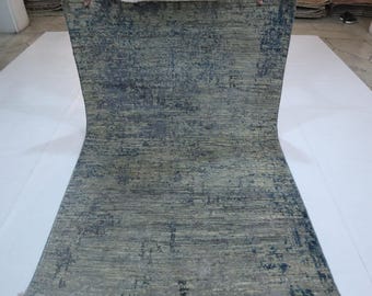 Puede incluir: Una alfombra rectangular con una apariencia desgastada y texturizada. La alfombra presenta una mezcla de tonos azul y beige apagados, creando una estética sutil y envejecida. El diseño tiene un efecto moteado, con acentos azules más oscuros. La alfombra está enrollada, mostrando todo su ancho.