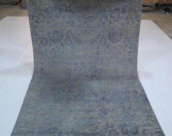 Puede incluir: Una alfombra rectangular enrollada con un diseño floral azul y gris descolorido. El diseño de la alfombra presenta un medallón central y motivos florales repetidos. La alfombra se exhibe sobre una superficie blanca.
