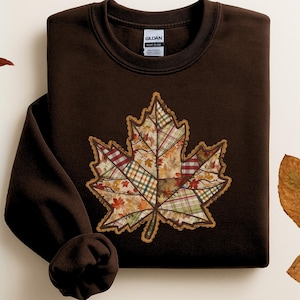 May include: Dark chocolate-coloured sweatshirt with a large, patchwork maple leaf design. The leaf is filled with various autumn-themed patterns and colours, including plaid and floral prints. The sweatshirt has a crew neck and long sleeves.