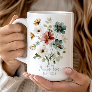 May include: White ceramic mug, 15 oz, decorated with a floral arrangement in shades of red, white, and teal. The mug is inscribed with "Auntie Tess" and "Est. 2026". The text "Graphic Never Fade" is at the top.
