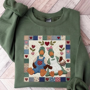 Puede incluir: Sudadera verde militar con un diseño acolchado con dos patos de dibujos animados. Uno lleva un peto, el otro un vestido y un delantal. El diseño incluye corazones y detalles florales, sobre un borde de patchwork.