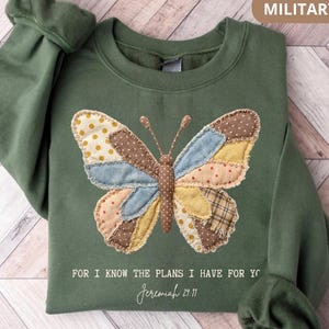 Peut inclure: Sweat-shirt vert militaire avec un motif de papillon en patchwork marron, bleu et jaune. Le texte "FOR I KNOW THE PLANS I HAVE FOR YOU" et "Jeremiah 29:11" est imprimé en dessous. Le sweat-shirt a des manches longues.