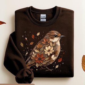 May include: A dark chocolate brown crewneck sweatshirt featuring a graphic of a bird with feathers composed of autumn-colored flowers and leaves. The Gildan brand label is visible at the inner neckline. The design includes scattered falling leaves.