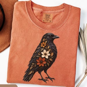 May include: A yam-colored t-shirt featuring a graphic of a black crow adorned with orange, white, and brown flowers on its chest. The shirt has a crew neckline and a Comfort Colors brand label printed on the inside of the collar in size small.