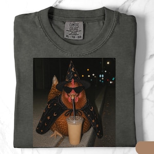 May include: A charcoal gray t-shirt featuring a graphic print of a brown chicken wearing a black witch hat, black sunglasses, and a black cape with orange star and moon patterns, while drinking from a clear plastic cup with a black straw.