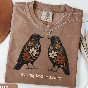 May include: An espresso-colored t-shirt featuring a graphic of two black birds with floral patterns on their chests. The text "attempted murder" is printed in white below the birds. The shirt has a Comfort Colors label showing size S-P-CH.