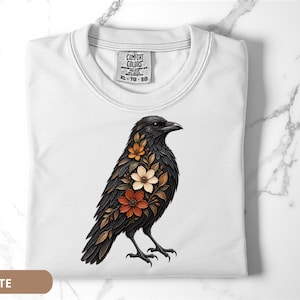 May include: A white t-shirt featuring a graphic of a black crow adorned with orange, brown, and cream-colored floral patterns on its chest. The shirt has a crew neckline and a Comfort Colors brand label printed on the inside of the collar area.
