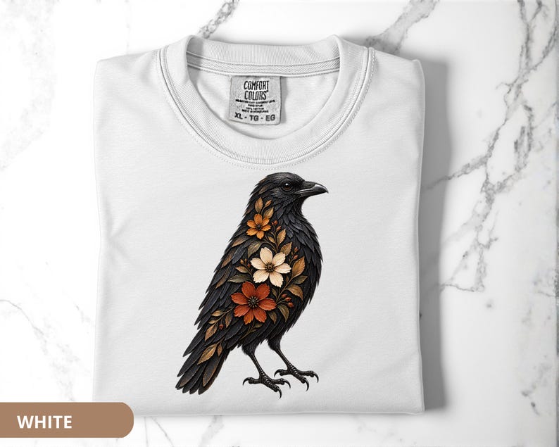 May include: A white t-shirt featuring a graphic of a black crow adorned with orange, brown, and cream-colored floral patterns on its chest. The shirt has a crew neckline and a Comfort Colors brand label printed on the inside of the collar area.