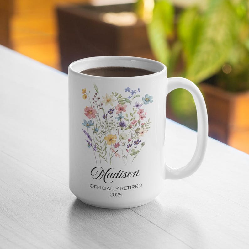 Op de afbeelding: Witte keramische mok met een bloemenontwerp in pastelkleuren, waaronder roze, blauw, geel en paars. De mok heeft de naam "Madison" en de tekst "Officially Retired 2025" in zwart.