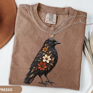 May include: A brown crew neck t-shirt featuring a graphic of a black crow adorned with orange, white, and brown flowers on its chest. The shirt has a Comfort Colors label at the neck, size S, and is displayed with a silver necklace and a hat.