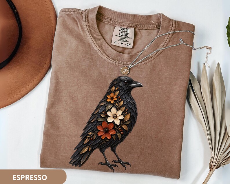 May include: A brown crew neck t-shirt featuring a graphic of a black crow adorned with orange, white, and brown flowers on its chest. The shirt has a Comfort Colors label at the neck, size S, and is displayed with a silver necklace and a hat.