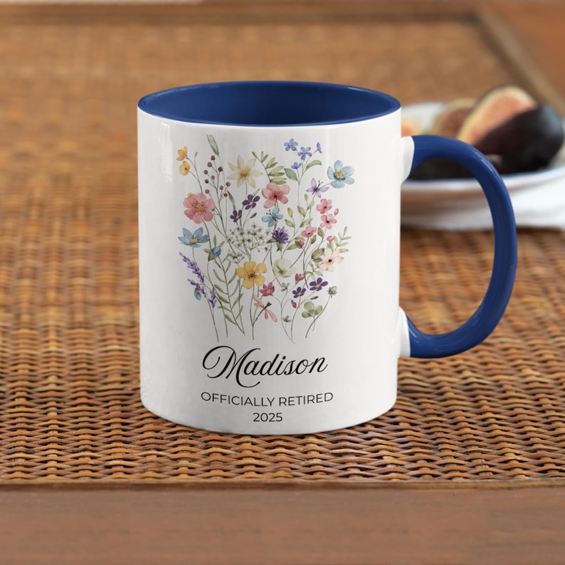 Op de afbeelding: Witte keramische mok met een blauw interieur en handvat, met een bloemenontwerp in pastelkleuren. De mok is gepersonaliseerd met de naam "Madison" en de tekst "OFFICIALLY RETIRED 2025".