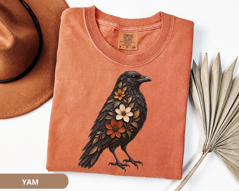 Floral raven shirt featuring a black raven decorated with cream, rust and burnt orange flowers on a Yam Comfort Colors tee, a gothic botanical bird design inspired by dark academia, dark cottagecore and witchy nature.