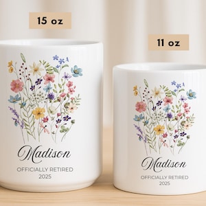 Op de afbeelding: Twee witte keramische mokken met bloemmotieven. De grotere mok, met het label 15 oz, heeft een boeket kleurrijke bloemen en de naam "Madison" met "OFFICIALLY RETIRED 2025". De kleinere mok, met het label 11 oz, heeft een vergelijkbaar ontwerp.