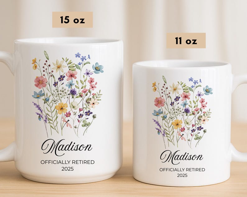 Op de afbeelding: Twee witte keramische mokken met bloemmotieven. De grotere mok, met het label 15 oz, heeft een boeket kleurrijke bloemen en de naam "Madison" met "OFFICIALLY RETIRED 2025". De kleinere mok, met het label 11 oz, heeft een vergelijkbaar ontwerp.