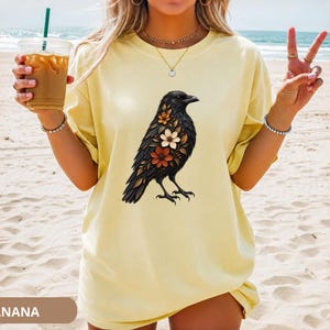 May include: A pale yellow short-sleeve t-shirt featuring a graphic print of a black crow adorned with various colorful flowers on its chest. The shirt has a crew neckline and a loose fit, showcasing a detailed illustration of the bird.