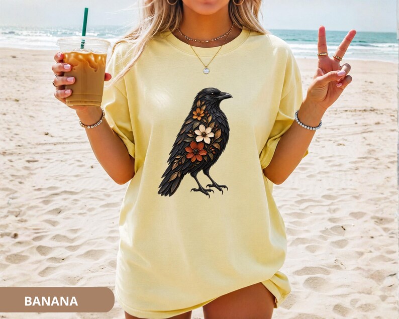 May include: A pale yellow short-sleeve t-shirt featuring a graphic print of a black crow adorned with various colorful flowers on its chest. The shirt has a crew neckline and a loose fit, showcasing a detailed illustration of the bird.