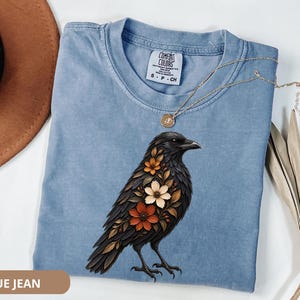 May include: A blue jean colored t-shirt featuring a graphic of a black crow with orange and cream colored flowers integrated into its plumage. The shirt has a Comfort Colors brand label at the neckline and is displayed with a gold necklace.