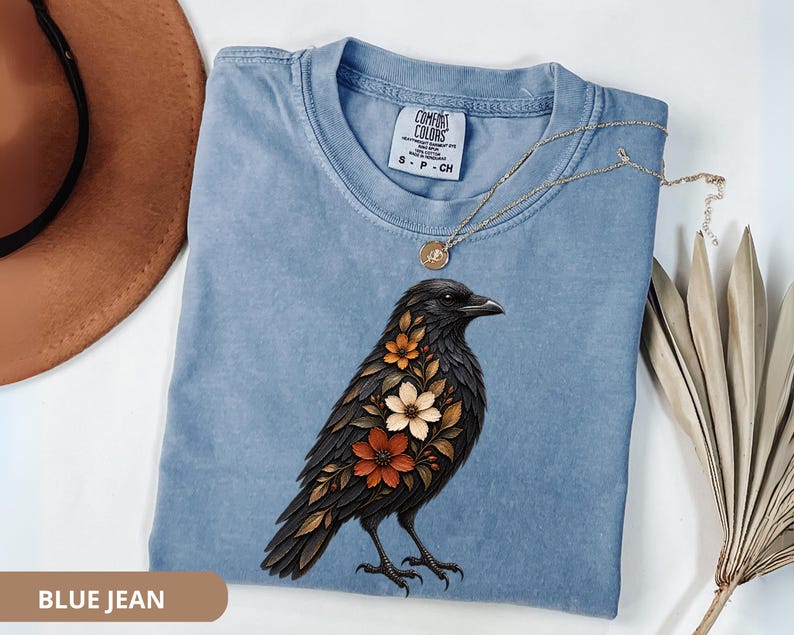 May include: A blue jean colored t-shirt featuring a graphic of a black crow with orange and cream colored flowers integrated into its plumage. The shirt has a Comfort Colors brand label at the neckline and is displayed with a gold necklace.