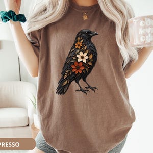 May include: A brown t-shirt featuring a graphic illustration of a black crow with orange, white, and brown flowers integrated into its plumage. The bird is depicted in profile, facing right, with detailed feathers and a floral pattern on its chest.