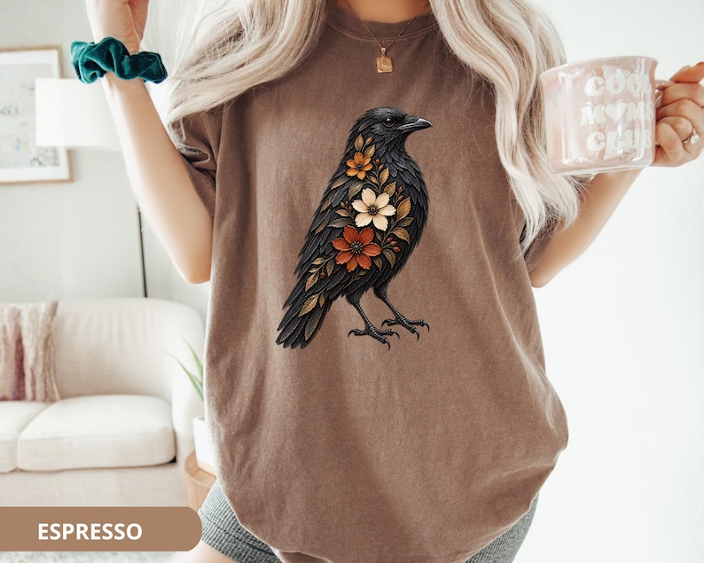 May include: A brown t-shirt featuring a graphic illustration of a black crow with orange, white, and brown flowers integrated into its plumage. The bird is depicted in profile, facing right, with detailed feathers and a floral pattern on its chest.