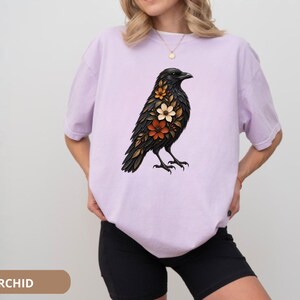 May include: A light purple short-sleeved t-shirt featuring a graphic print of a black crow adorned with orange, white, and brown flowers on its chest. The shirt has a crew neckline and a loose, relaxed fit, showcasing a detailed bird illustration.