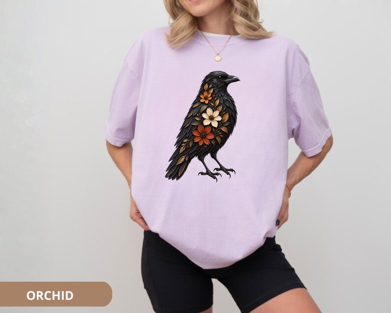 May include: A light purple short-sleeved t-shirt featuring a graphic print of a black crow adorned with orange, white, and brown flowers on its chest. The shirt has a crew neckline and a loose, relaxed fit, showcasing a detailed bird illustration.