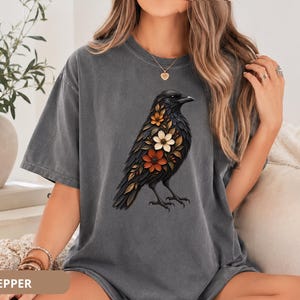 May include: A charcoal gray short-sleeve t-shirt featuring a graphic print of a black crow with orange, white, and brown flowers integrated into its plumage. The shirt has a crew neckline and a relaxed fit, showcasing a detailed bird illustration.