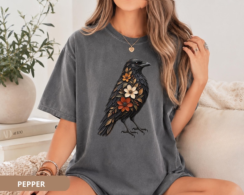 May include: A charcoal gray short-sleeve t-shirt featuring a graphic print of a black crow with orange, white, and brown flowers integrated into its plumage. The shirt has a crew neckline and a relaxed fit, showcasing a detailed bird illustration.