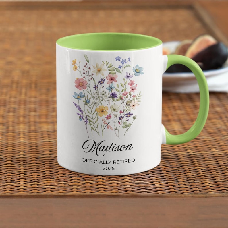 Op de afbeelding: Witte keramische mok met een groene binnenkant en handvat. De mok heeft een bloemenontwerp met diverse kleurrijke bloemen. De naam "Madison" staat in script, met "OFFICIALLY RETIRED 2025" eronder.
