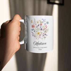 Op de afbeelding: Witte keramische mok met een bloemenontwerp met roze, gele, blauwe en paarse bloemen. De mok heeft de naam "Madison" en de tekst "OFFICIALLY RETIRED 2025" in zwart schrift.
