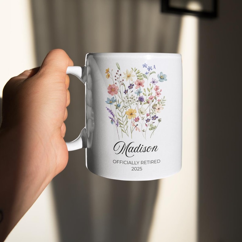 Op de afbeelding: Witte keramische mok met een bloemenontwerp met roze, gele, blauwe en paarse bloemen. De mok heeft de naam "Madison" en de tekst "OFFICIALLY RETIRED 2025" in zwart schrift.