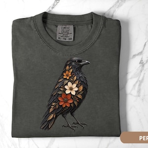 May include: A charcoal gray t-shirt featuring a graphic of a black crow adorned with orange, cream, and brown floral patterns on its chest. The shirt has a crew neckline and a Comfort Colors brand label printed on the inside of the collar.
