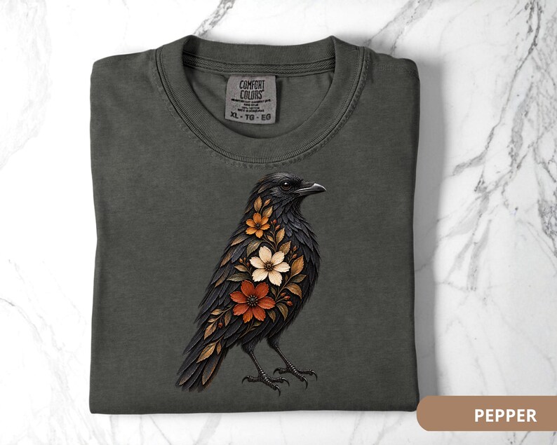 May include: A charcoal gray t-shirt featuring a graphic of a black crow adorned with orange, cream, and brown floral patterns on its chest. The shirt has a crew neckline and a Comfort Colors brand label printed on the inside of the collar.