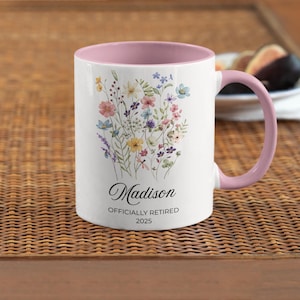 Op de afbeelding: Witte keramische mok met een roze binnenkant en handvat. De mok heeft een bloemenboeketontwerp in pastelkleuren en de naam "Madison" met de tekst "OFFICIALLY RETIRED 2025".
